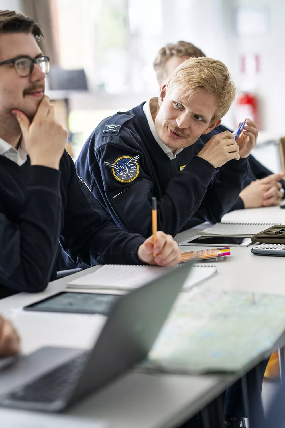 Glad manlig pilotstuderande i uniform sitter i klassrum, vänder sig mot en kompis. Foto.