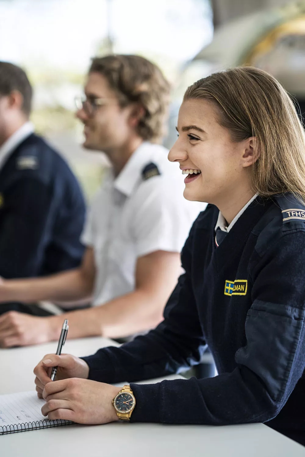 Leende kvinnlig pilotstuderande i unifrom sitter vid bänk i klassrum. Foto.