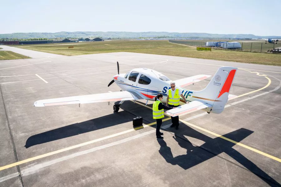 Kvinnlig pilotstuderande och manlig flyginstruktör står vid flygplan på landningsbana. Foto.